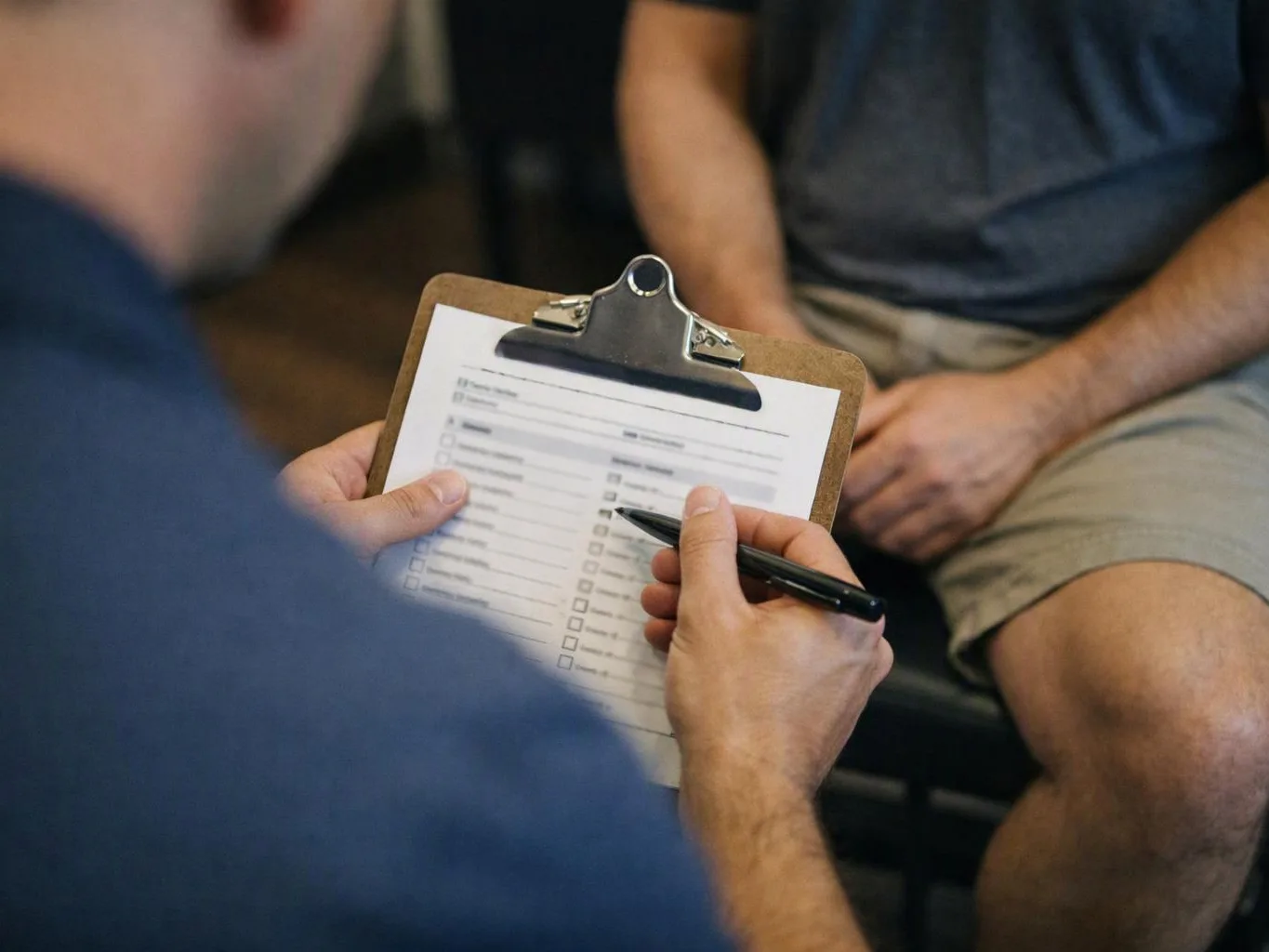 A person fills out a checklist on a clipboard while another person sits nearby in a room